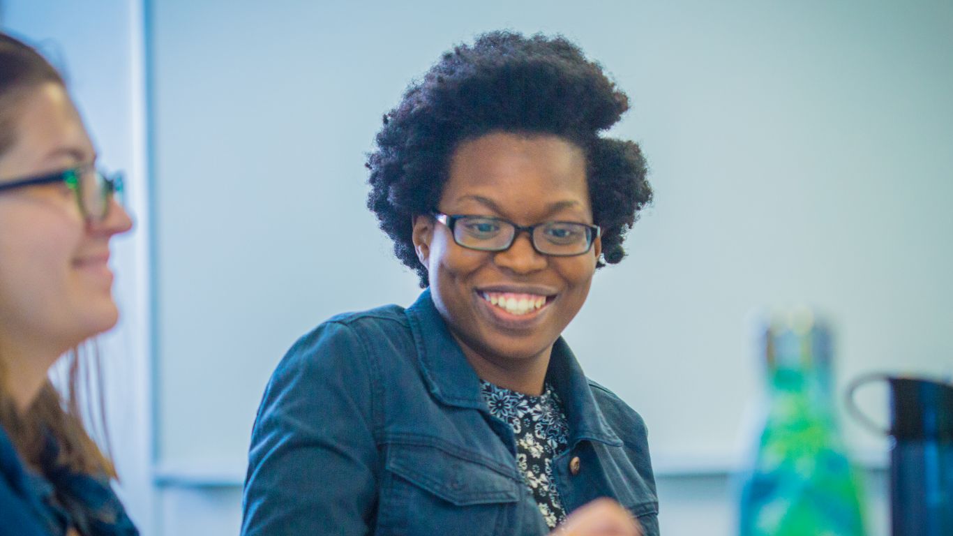 A smiling woman with curly hair sits at a table during a meeting, engaging with colleagues.