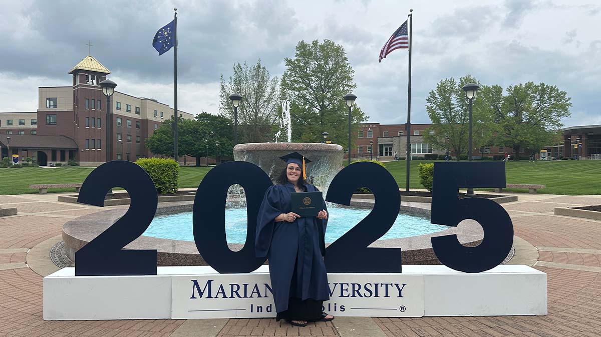 A graduation photo of a student posing with a diploma in front of a large "2025" sign at Marian University, surrounded by a scenic campus backdrop.