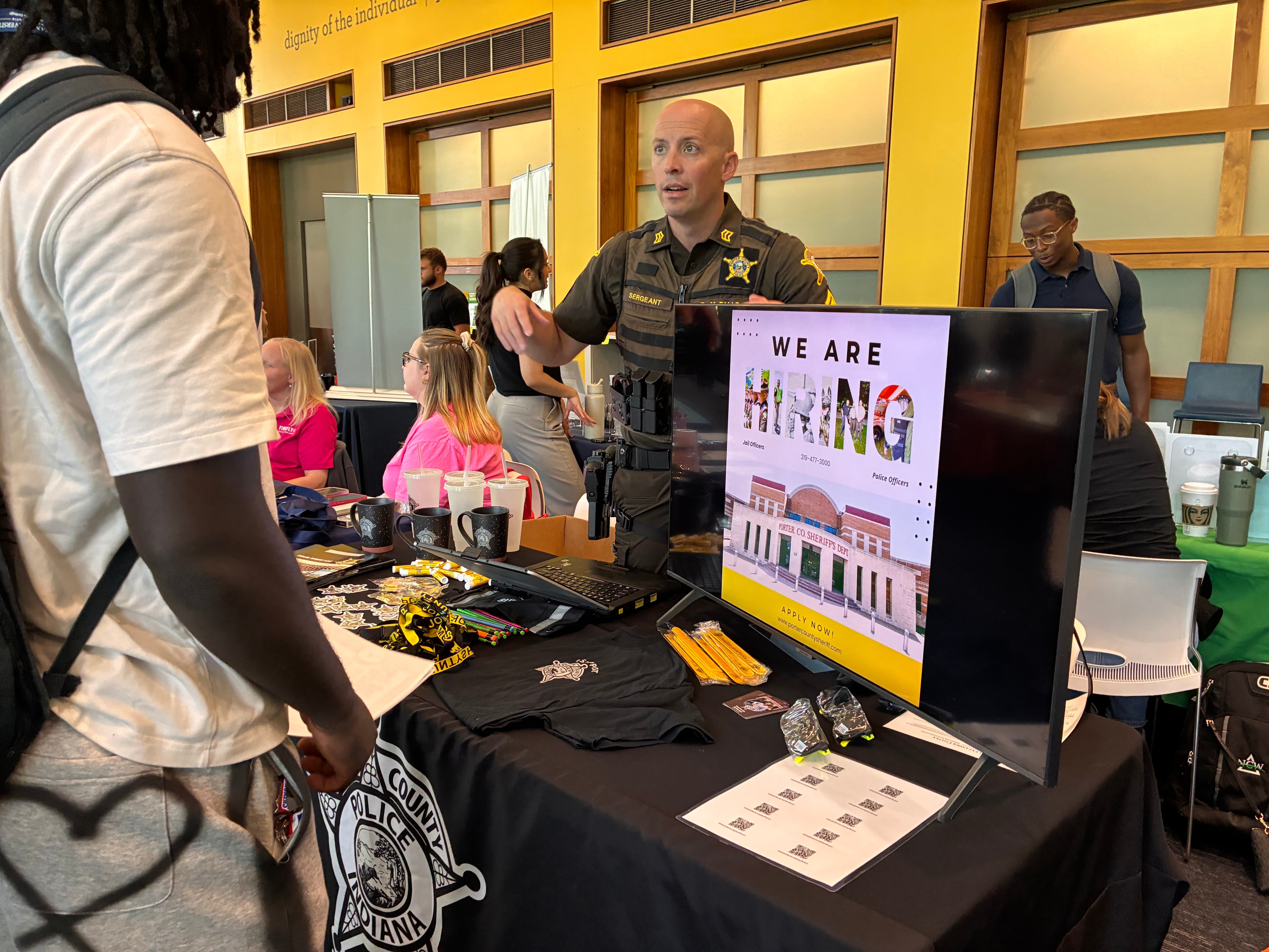 A law enforcement representative at a recruitment booth engaging with a potential applicant, featuring a large sign that says "WE ARE HIRING."