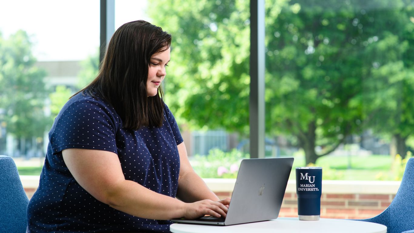 A college student working on a laptop at a bright, open study space, with a Marian University coffee cup on the table.