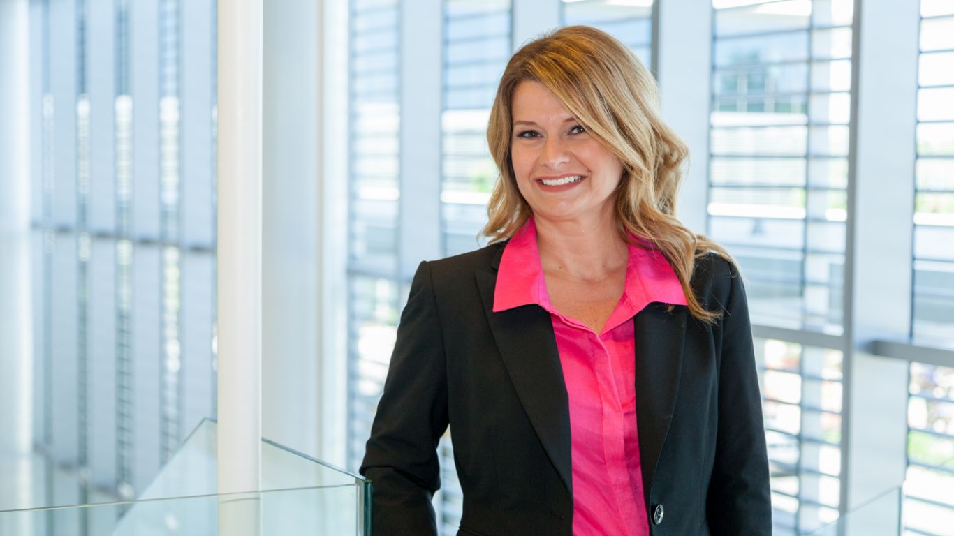 Professional headshot of a woman in a black blazer and pink shirt, standing in a modern office environment with large windows.